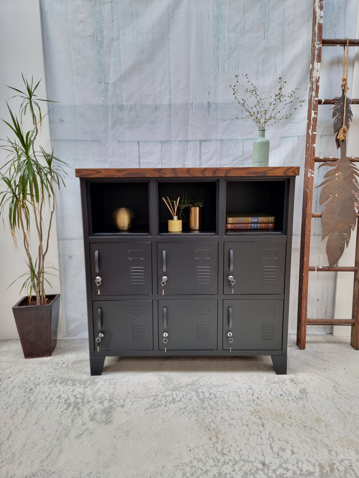Black metal 9-door locker with dark oak top, featuring a potted plant to the left and a ladder in the background.