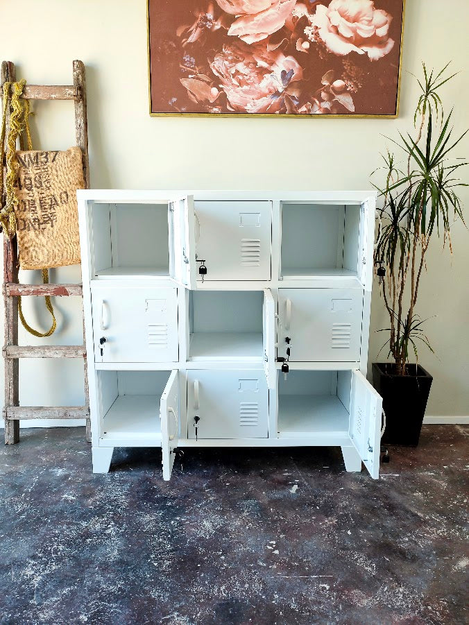 A white 9-compartment metal locker cabinet with doors on some compartments, shown in a room setting with a rustic wooden ladder, a potted plant, and a floral painting on the wall.