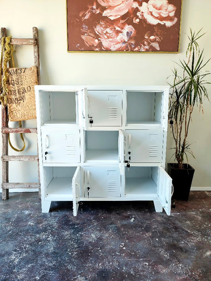 A white 9-compartment metal locker cabinet with doors on some compartments, shown in a room setting with a rustic wooden ladder, a potted plant, and a floral painting on the wall.