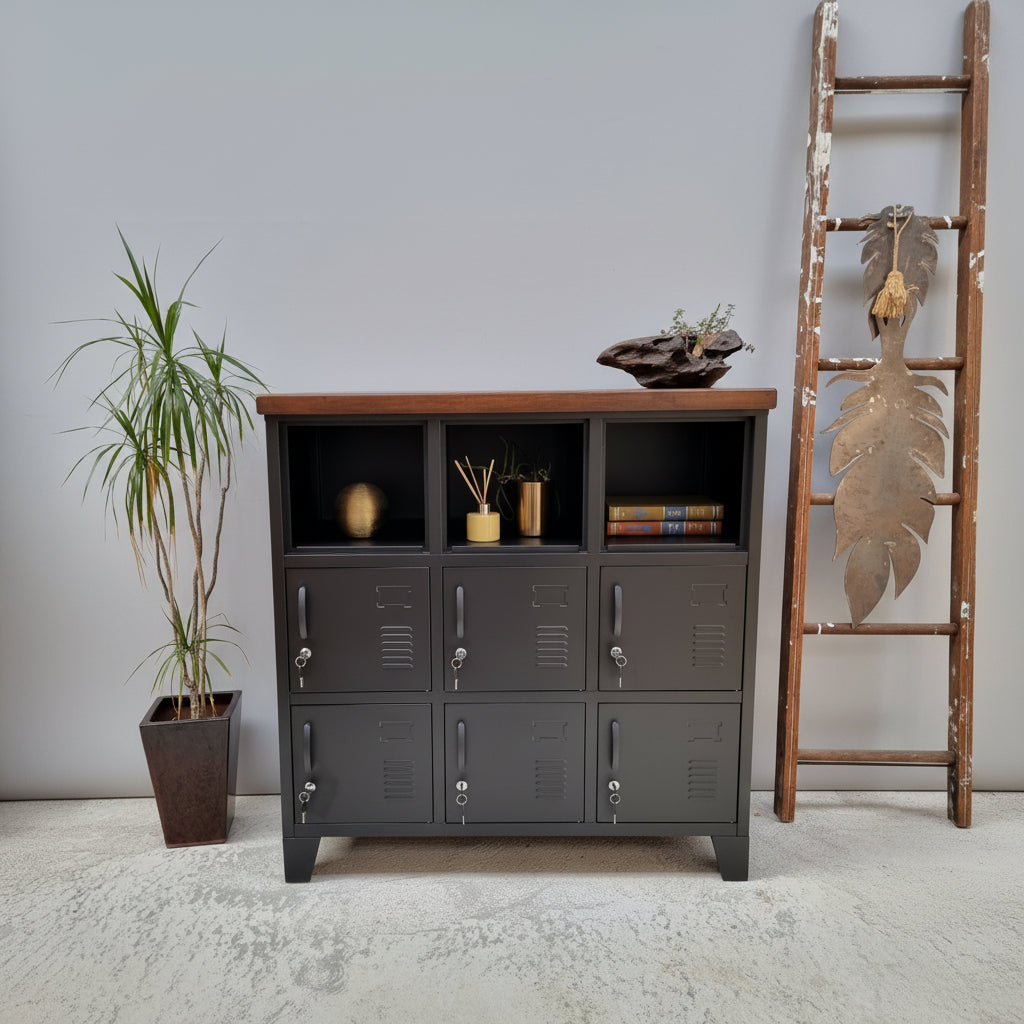 A black 9-door metal locker with a dark Rimu top, situated against a wall with a ladder and a potted plant beside it. The locker has a industrial style with visible handles and a textured finish.