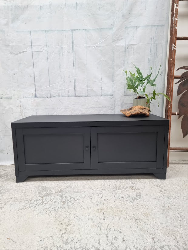 Black industrial metal storage bench with a plant and blanket against a light gray wall.