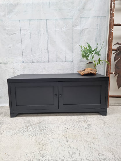 Black industrial metal storage bench with a plant and blanket against a light gray wall.