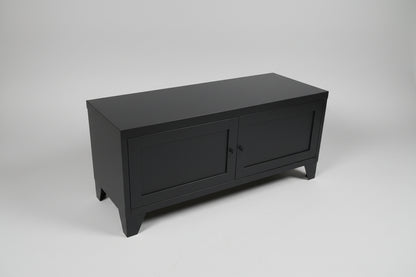 Black metal TV cabinet with two paneled doors on a textured floor with a plant in the background