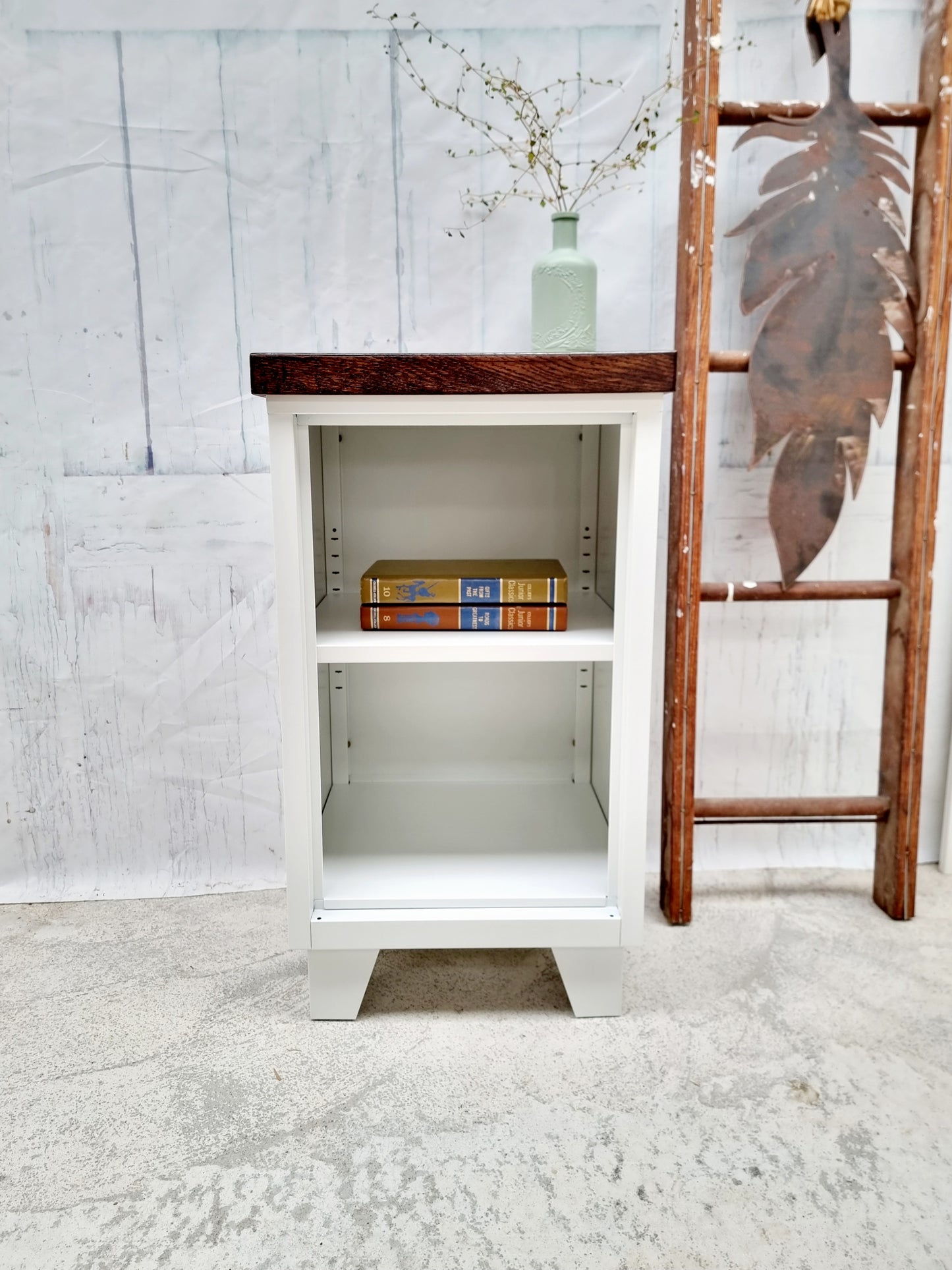 White metal cabinet with wooden top and sides, containing books and a vase, against a light gray wall.