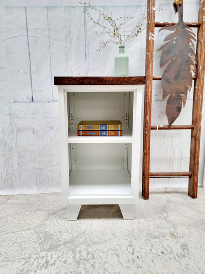 White metal cabinet with wooden top and sides, containing books and a vase, against a light gray wall.
