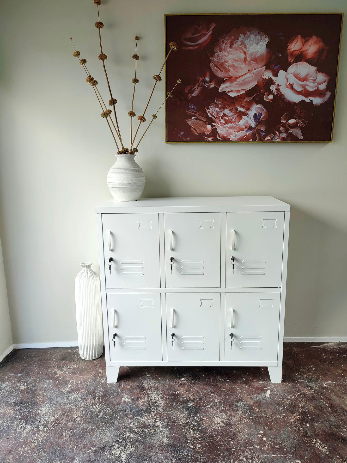 A white 6-door metal locker with each door featuring a key lock. It is positioned against a wall with a floral artwork above it, alongside a white vase with dried flowers.