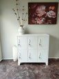 A white 6-door metal locker with each door featuring a key lock. It is positioned against a wall with a floral artwork above it, alongside a white vase with dried flowers.
