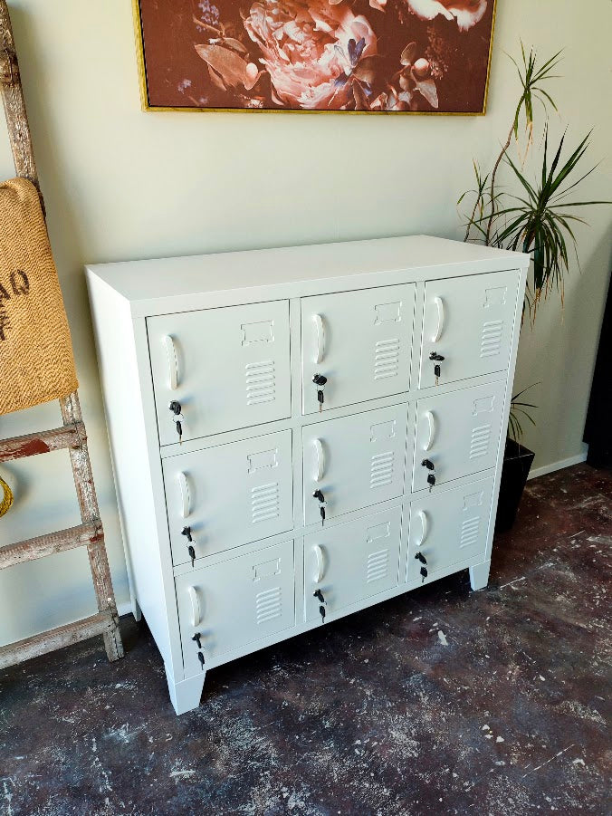 A white metal storage cabinet with nine lockable doors, styled in a room setting with a dark patterned floor, a potted plant, and a rustic wooden ladder.