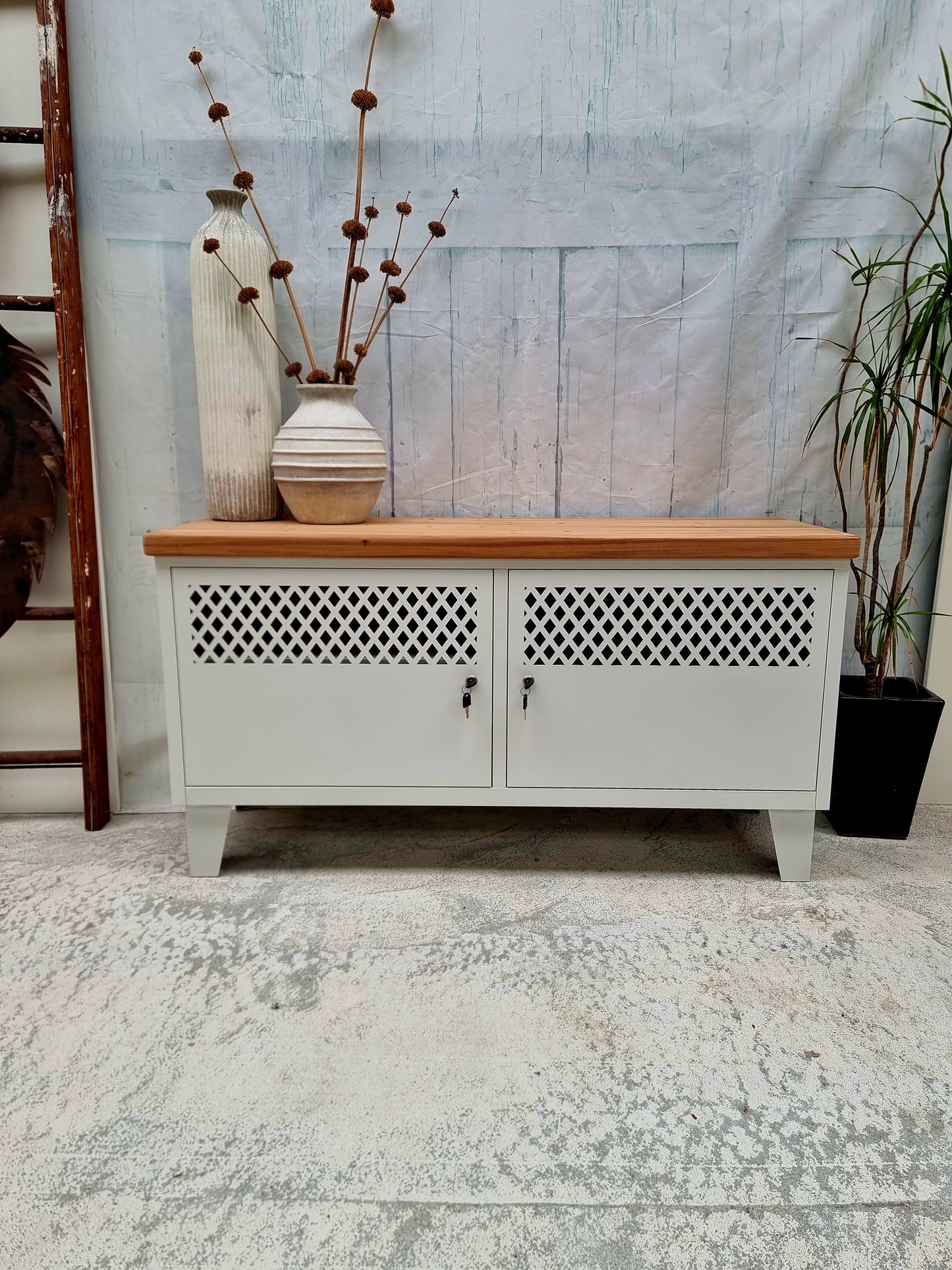 A white metal industrial TV cabinet with dark stained recycled Rimu top, featuring two doors with metal grid designs, set against a distressed wooden backdrop.