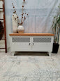 A white metal industrial TV cabinet with dark stained recycled Rimu top, featuring two doors with metal grid designs, set against a distressed wooden backdrop.