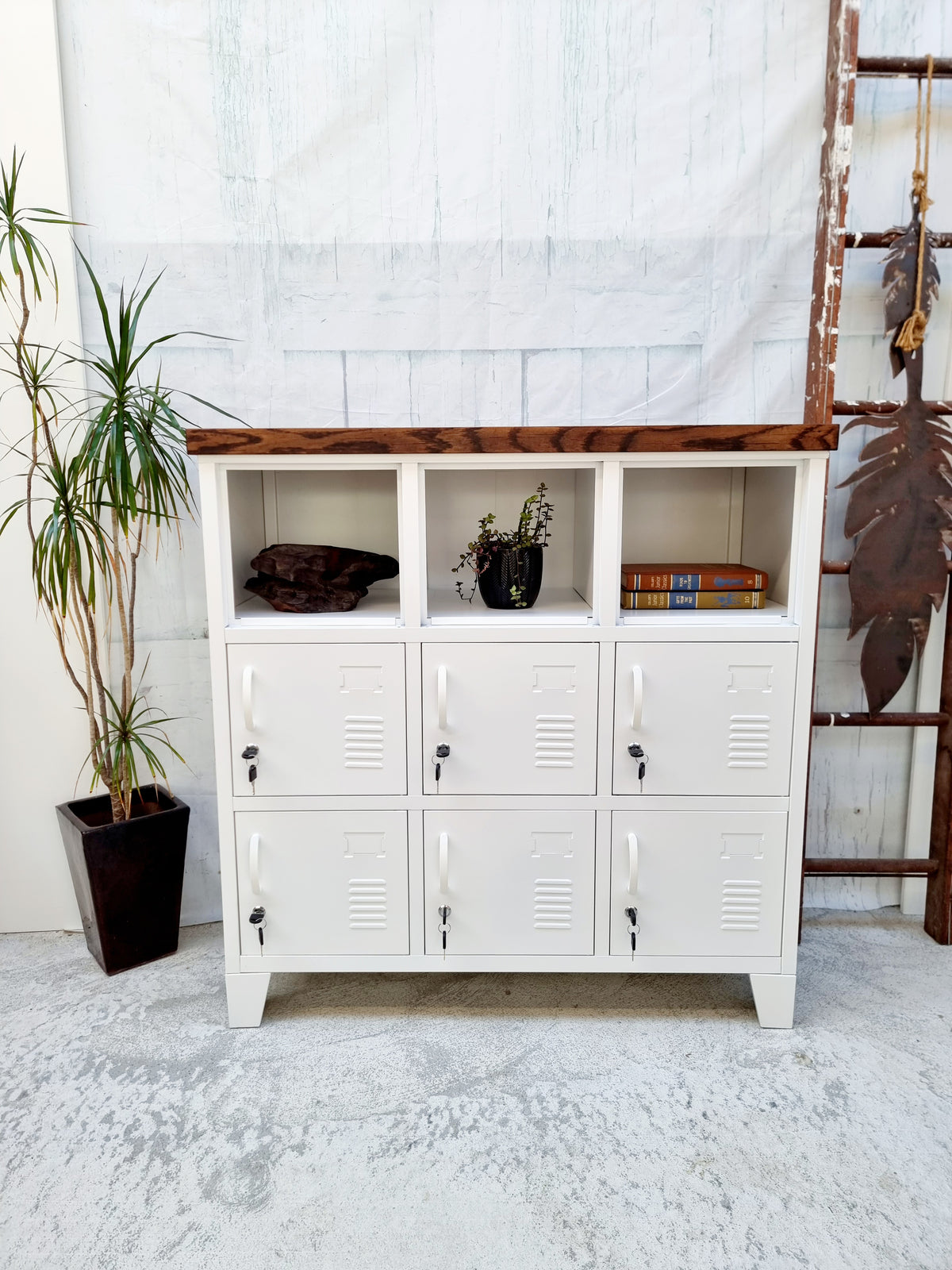 A white 9-door locker with a dark oak top, situated against a white background with a potted plant to the side and a decorative item on top of the locker.