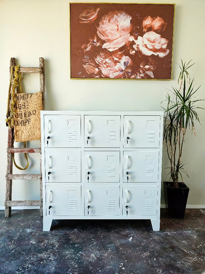 A white 9-door metal locker cabinet stands against a beige wall in a styled room setting, with a floral painting above, a rustic ladder on the left, and a potted plant on the right.