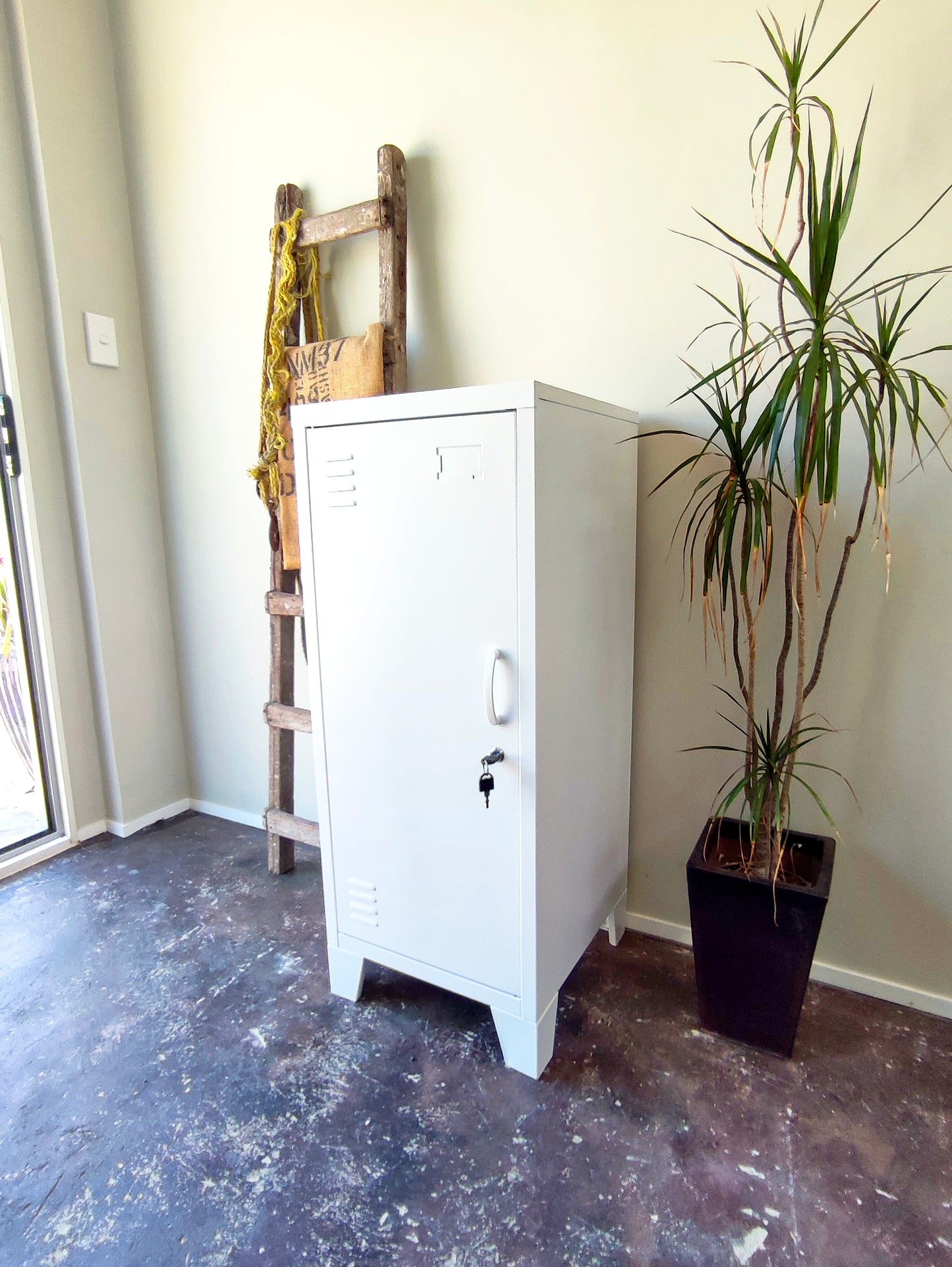 A white metal side cabinet with a left hinge, two adjustable shelves, and a lock on the door. It is placed in a room next to a potted plant and a wooden ladder.