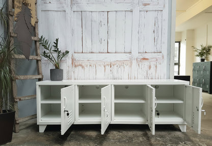 Large White metal entertainment cabinet with open doors showing cable holes and shelves inside in a room with wooden walls and plants.