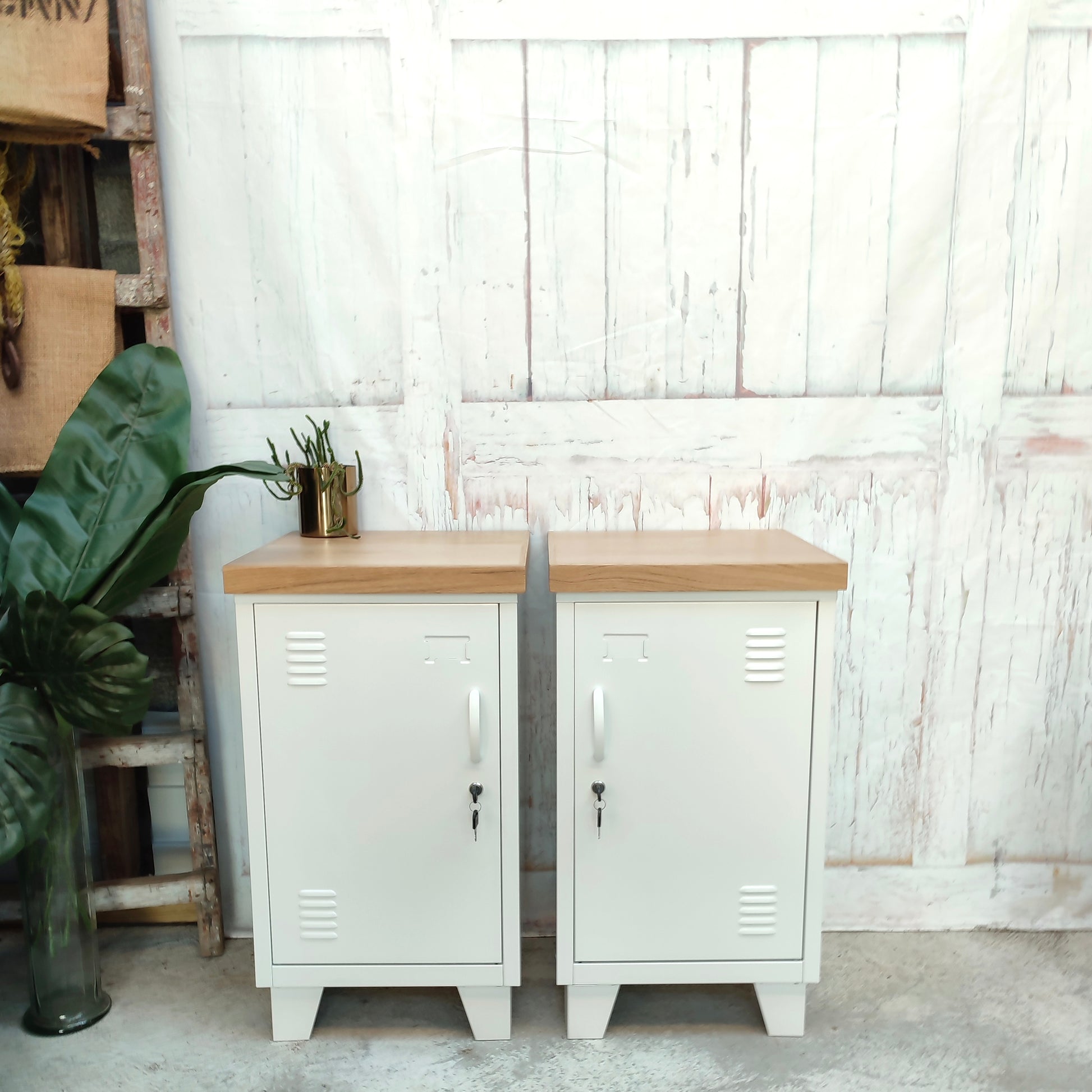 A pair of white mini lockers with pale oak tops, featuring lock mechanisms, set against a rustic backdrop with a plant to the side.