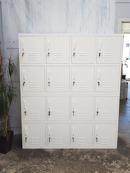 A white metal 16-door locker unit with vertical vents on each door, situated against a wall with a plant to its side.
