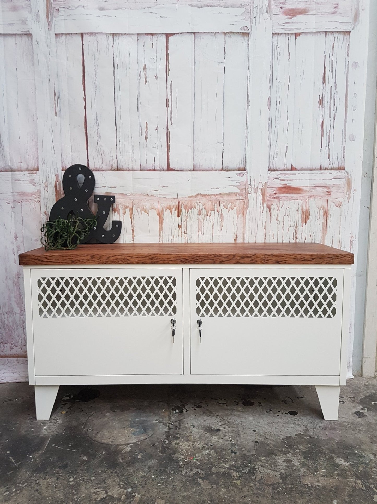 A white TV cabinet with dark oak top featuring metal grid design on the doors, set against a rustic backdrop.