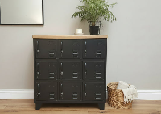 A black metal 9-door locker with an oak top, positioned against a white wooden background with a green potted plant to the side.