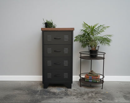 A black metal 4-drawer cabinet with a dark oak top, positioned in a room with a plant and a round metal side table beside it.