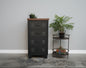 A black metal 4-drawer cabinet with a dark oak top, positioned in a room with a plant and a round metal side table beside it.