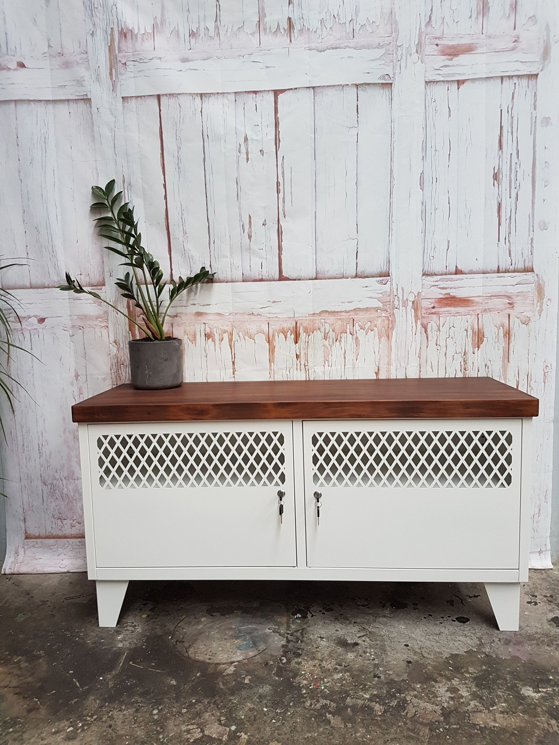 A white metal industrial TV cabinet with dark stained recycled Rimu top, featuring two doors with metal grid designs, set against a distressed wooden backdrop.